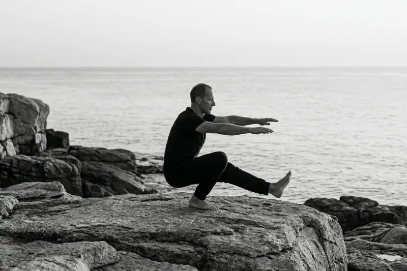 Ein erfahrener Athlet führt regelmäßig Sport treibend eine kontrollierte Einbein-Kniebeuge auf einem Felsen am Meer aus, symbolisiert Souveränität, Autarkie und das KISS-Prinzip.