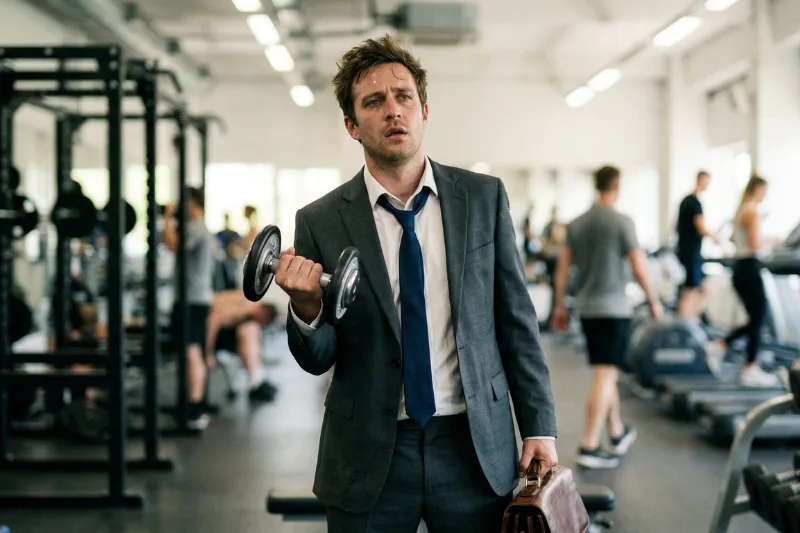 Ein sichtlich gestresster Mann im grauen Anzug mit gelockerter Krawatte steht verloren in einem modernen Fitnessstudio. Er hält in der einen Hand eine schwere Aktentasche und macht mit der anderen Hand einen Hantel-Curl, während er erschöpft ins Leere blickt.