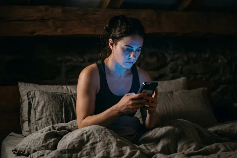 Eine sichtlich müde, erschöpfte Frau sitzt spät in der Nacht im Bett und starrt auf ihr leuchtendes Smartphone-Display. Das kühle, künstliche Licht des Bildschirms beleuchtet ihr ernstes Gesicht in einem dunklen, rustikalen Schlafzimmer mit Steinwänden und Holzdeckenträgern. Dies visualisiert Schlafmangel und digitale Ablenkung, die Regeneration und Muskelaufbau im Keim ersticken.
