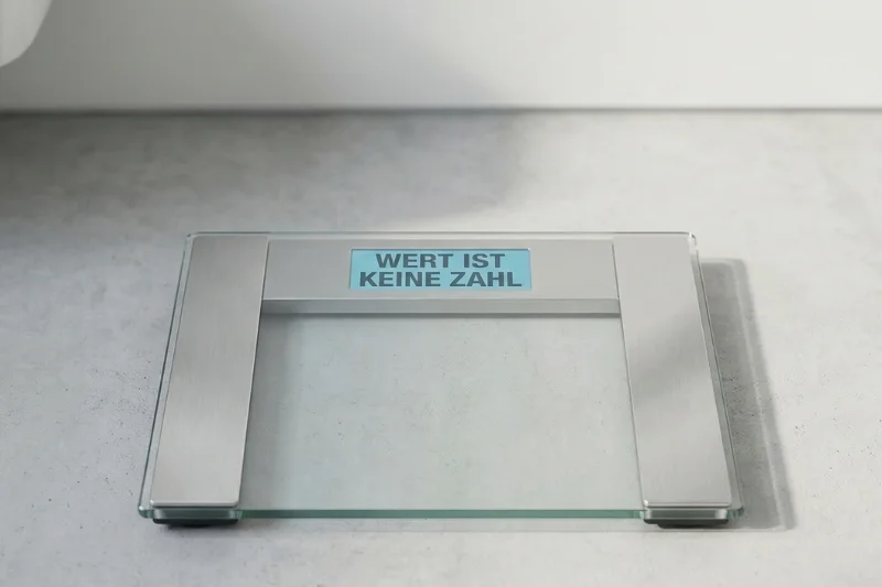 Eine moderne digitale Personenwaage auf grauem Boden, deren hellblau leuchtendes Display den Schriftzug „WERT IST KEINE ZAHL“ zeigt.