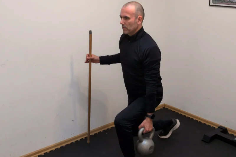 Ausfallschritte mit Unterstützung: Besenstiel in der einen Hand, eine Kettlebell in der anderen.