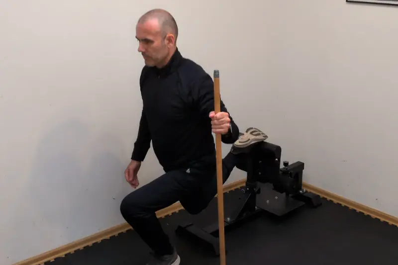 Bulgarian Split Squats mit Unterstützung: Ein Fuß auf einer Erhöhung abgesetzt, in der anderen Hand der Besenstiel.