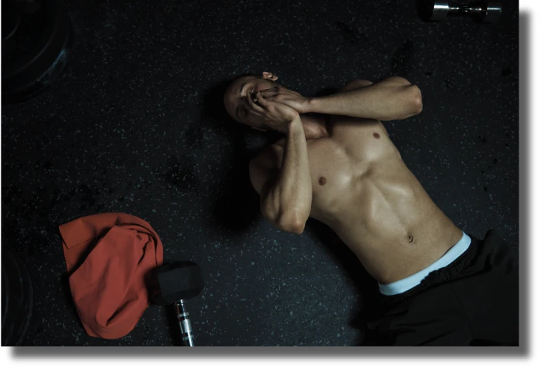 Ein erschöpfter trainierter Mann liegt auf dem Boden eines Fitnessstudios. Neben ihm liegt ein Kurzhantel. Symbolbild für den Kult der Erschöpfung im Training.